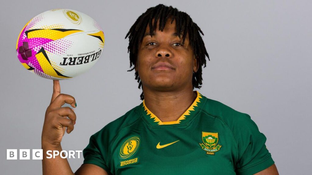 Babalwa Latsha, wearing a green South Africa rugby shirt in front of a grey background, looks directly at the camera as she balances a rugby ball on her left index finger