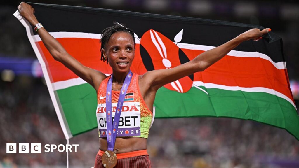 Beatrice Chebet wears a gold medal on a purple ribbon over an orange and green Kenya running vest as she holds a Kenyan flag aloft behind her. She has a smile on her face as she celebrates winning the 10,000m at the World Athletics Championships in Tokyo